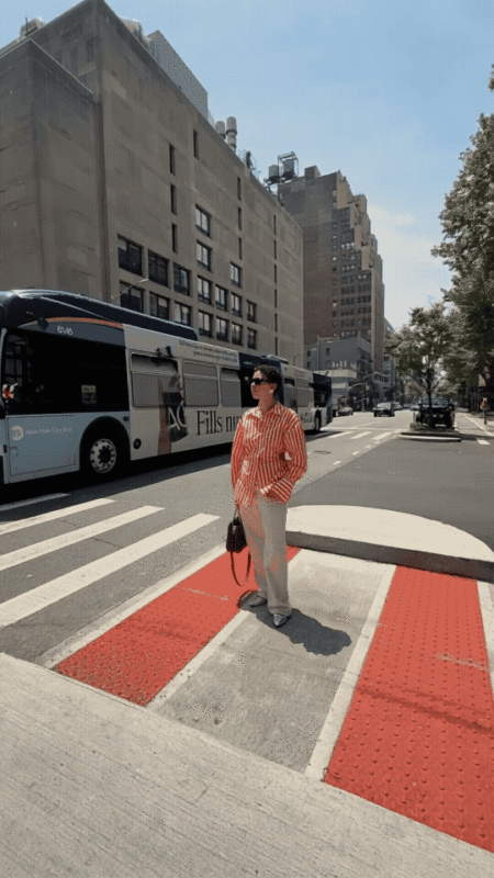 A person in a red and white striped shirt, white pants, and sunglasses stands at a crosswalk in a city. A bus and tall buildings are visible in the background under a clear sky.