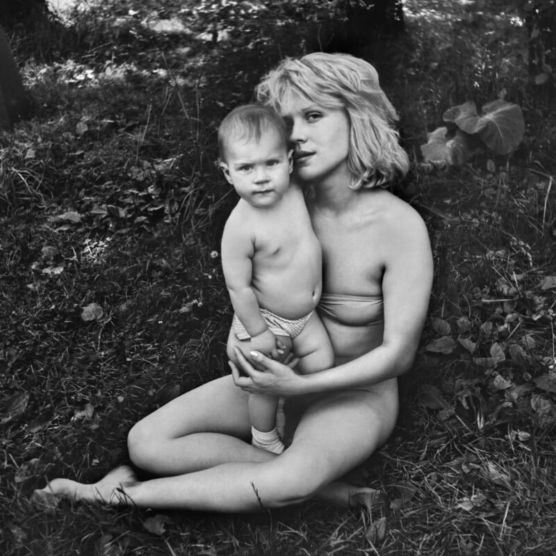 A woman in a bikini sits on grass outdoors, holding a young child who is standing and wearing only a diaper and socks. They are surrounded by leaves and plants, and both look directly at the camera.