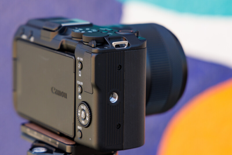 Close-up of a black Canon camera mounted on a tripod, focusing on the side buttons and lens. The background features blurred, colorful abstract shapes in blue, white, and orange.