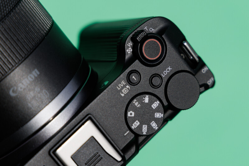 Close-up of a digital camera with its control dials and buttons visible. The camera is angled on a green background, highlighting the textured buttons and adjustment knobs around the shutter release button.