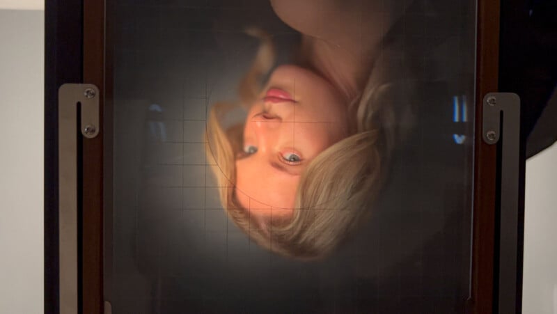 A blonde woman’s face is seen upside down and softly focused through the ground glass of a large format camera, framed by the camera’s edges and grid lines.