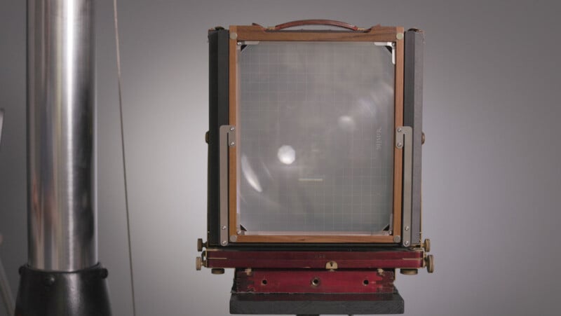 A large format vintage camera with a wooden frame and a ground glass focusing screen, shown front-facing on a tripod against a plain gray background.
