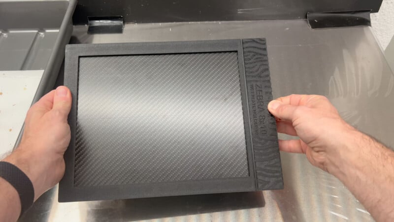 A person’s hands hold a black rectangular object labeled “ZEBRA 8×10,” featuring a textured carbon-fiber pattern, above a metallic industrial surface.