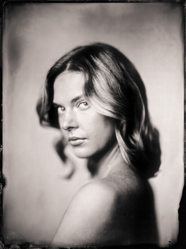 Black and white portrait of a person with shoulder-length wavy hair looking over their shoulder, gazing softly at the camera, with a blurred background and soft lighting.