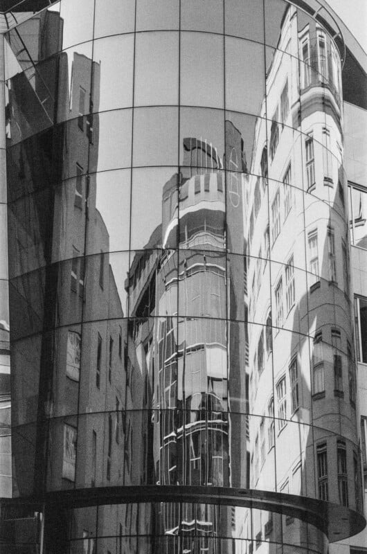 A modern glass building reflects distorted images of nearby older buildings, creating an abstract, wavy pattern across its mirrored surface. The scene is in black and white.