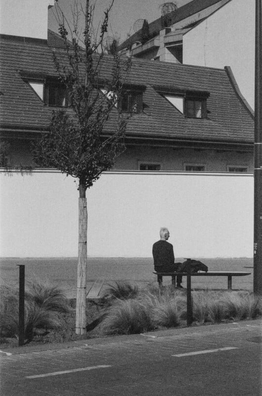 Black-and-white photo of a person sitting alone on a bench in front of a small tree, with a building in the background and a quiet street in the foreground.