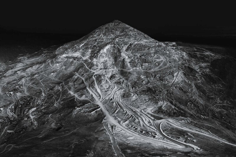 A dramatic, black and white aerial view of a mountain heavily scarred by winding mining roads and pathways, creating a stark, intricate pattern across its surface under a dark sky.