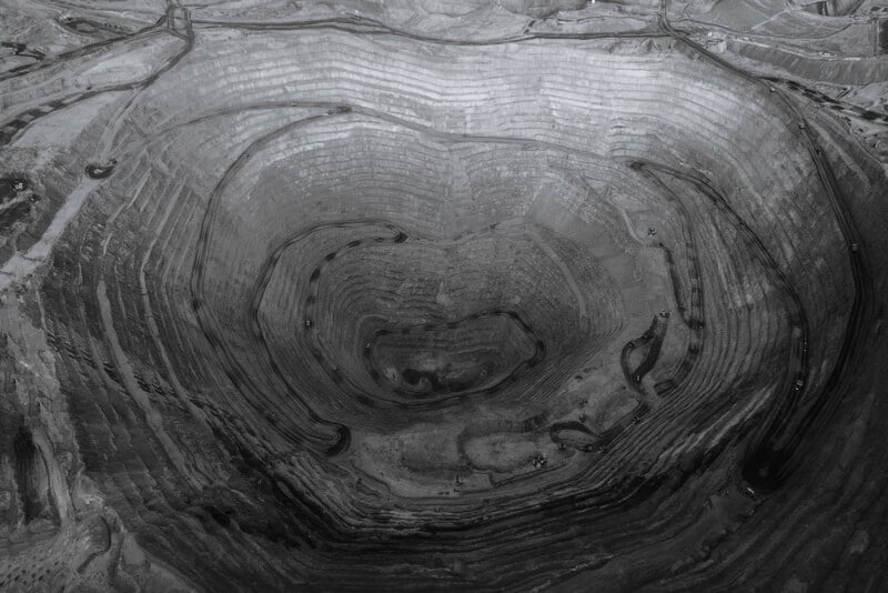 Aerial view of a large open-pit mine with terraced, concentric circular layers descending into the earth; vehicles and machinery are visible on some of the levels. The image is in black and white.
