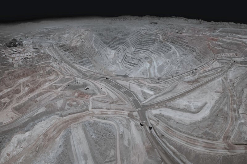 Aerial view of a large open-pit mine with terraced, stepped sides and winding dirt roads. Several mining vehicles are visible on the roads within the arid, dusty landscape. The sky is dark and overcast.