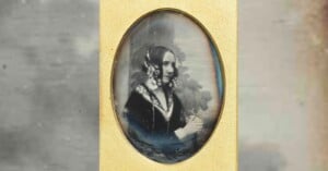 A sepia-toned portrait of a woman in Victorian-era clothing, with floral hair accessories, seated and looking to the side. The image is oval-shaped and framed in gold.
