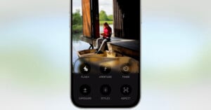 A smartphone screen displays a camera app interface with settings like flash, aperture, and timer. Behind the interface, a woman sits on a dock by the water, reading a book near an open wooden structure.