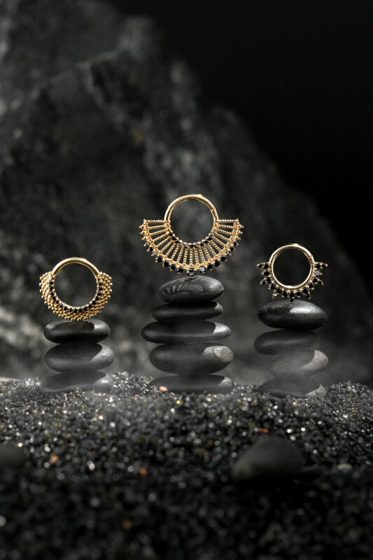 Three ornate gold rings are displayed on stacks of smooth black stones, with a misty, dark rock background creating a dramatic and elegant atmosphere.