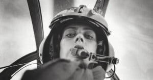 Black and white photo of a pilot wearing a helmet and headset, looking down into the camera from inside a cockpit, with blurred background and cockpit structures visible.