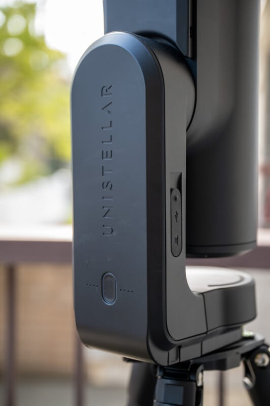 A close-up of a black device labeled "UNISTELLAR," mounted on a tripod outdoors, with a blurred background of trees and a building.