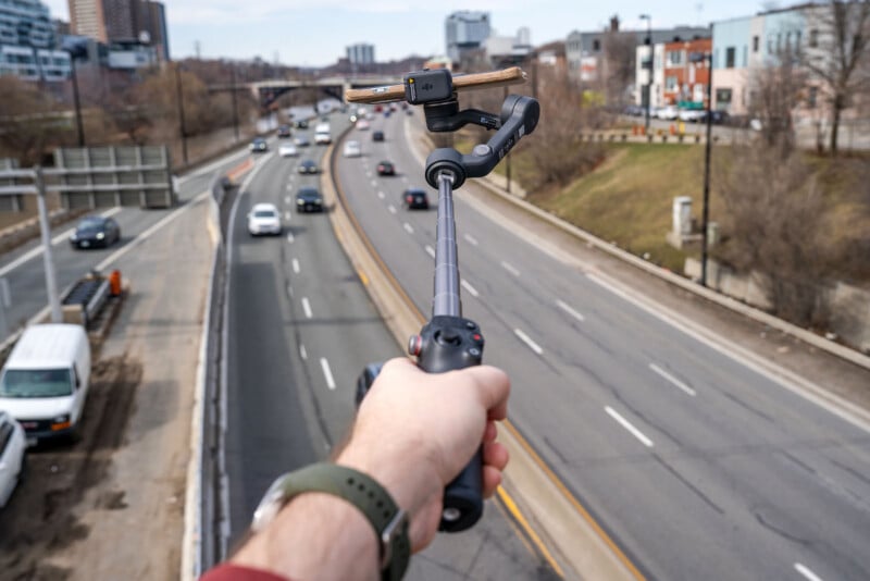 A person holds a selfie stick with a camera or phone mounted on it, extending over a busy multi-lane highway with cars, city buildings, and trees visible in the background.