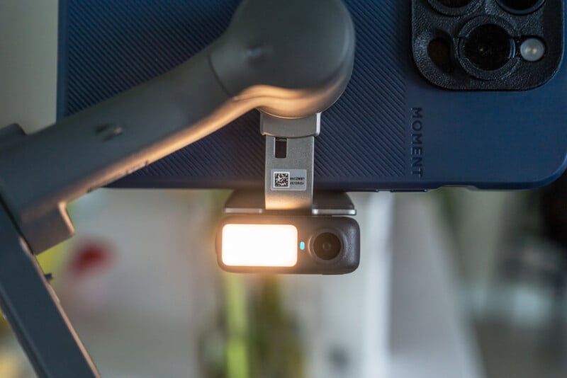 A close-up of a smartphone mounted on a gray gimbal stabilizer, with the phone's camera facing forward and a blurry background with light and colors.