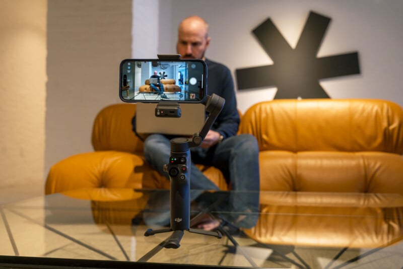 A smartphone mounted on a DJI gimbal records a man sitting on a yellow sofa with a black asterisk-shaped wall decoration behind him. The scene is shot through a glass table.