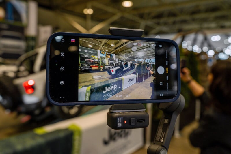 A smartphone mounted on a gimbal records a white Jeep vehicle at an indoor auto show, capturing the scene through its camera screen, with blurred people and banners in the background.