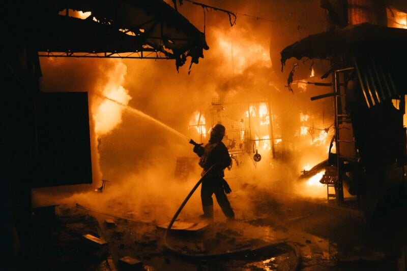 A firefighter sprays water on a blazing fire inside a building at night, surrounded by thick smoke and burning debris. The intense flames illuminate the scene with an orange glow.