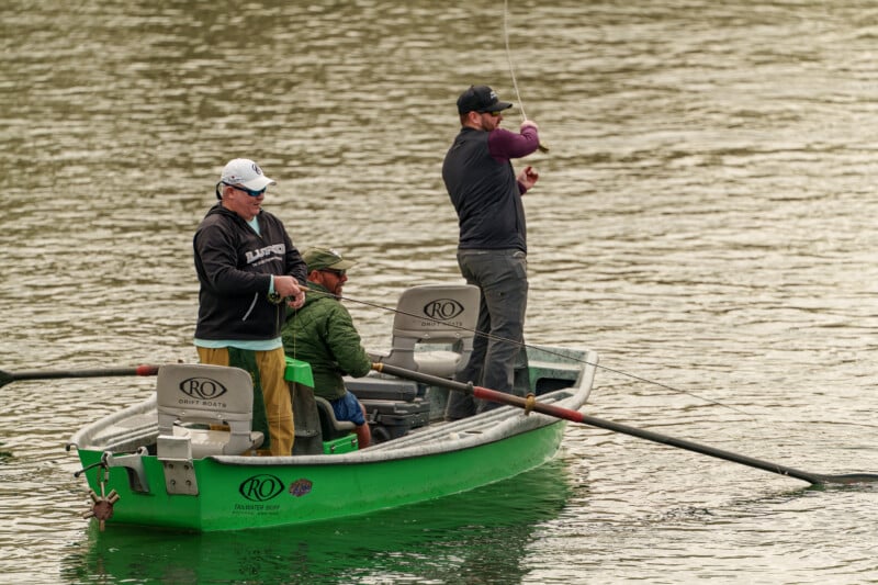 Three people are fishing from a small green boat on a calm body of water. One person is casting a line, another is seated, and the third is rowing. The scene appears relaxed and peaceful.