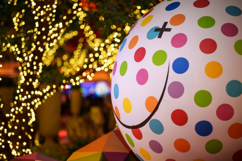 A large white sphere with colorful polka dots and a cartoon-style face with a black X for one eye and a curved smile, set against a background of twinkling yellow lights and blurred trees.
