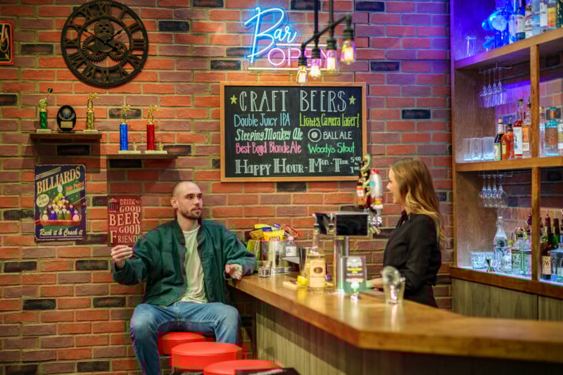 A man sits at a bar counter talking to a bartender in a cozy pub with brick walls, neon signs, trophies, beer taps, and colorful decor. A “Craft Beers” chalkboard menu is displayed on the wall behind them.