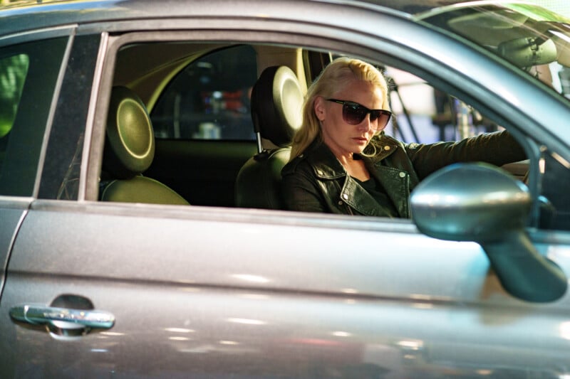 A woman wearing sunglasses and a black leather jacket sits in the driver's seat of a silver car, looking out the open window with a serious expression.