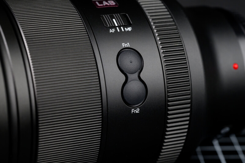 Close-up of a camera lens with visible focus modes labeled "AF" and "MF." Two round buttons labeled "Fn1" and "Fn2" are prominently displayed. The lens surface features textured grip rings and a small red dot for alignment.