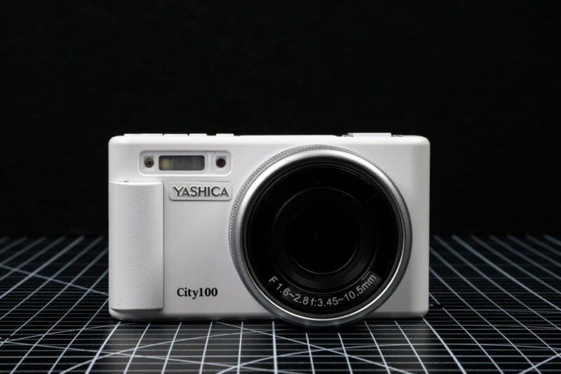 A white Yashica City100 digital camera is placed on a black surface with a grid pattern, against a dark background. The camera lens and brand name are clearly visible.
