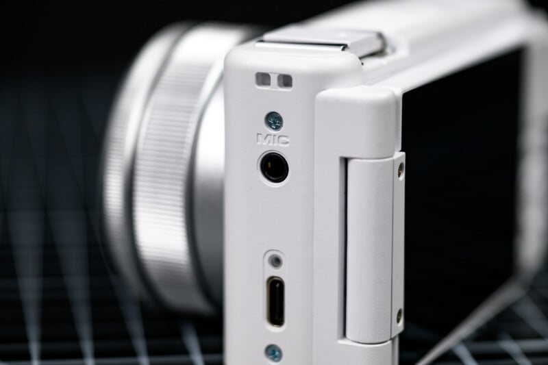 Close-up of the side of a white digital camera showing a microphone port, a USB-C port, and a hinge against a dark, grid-patterned background.