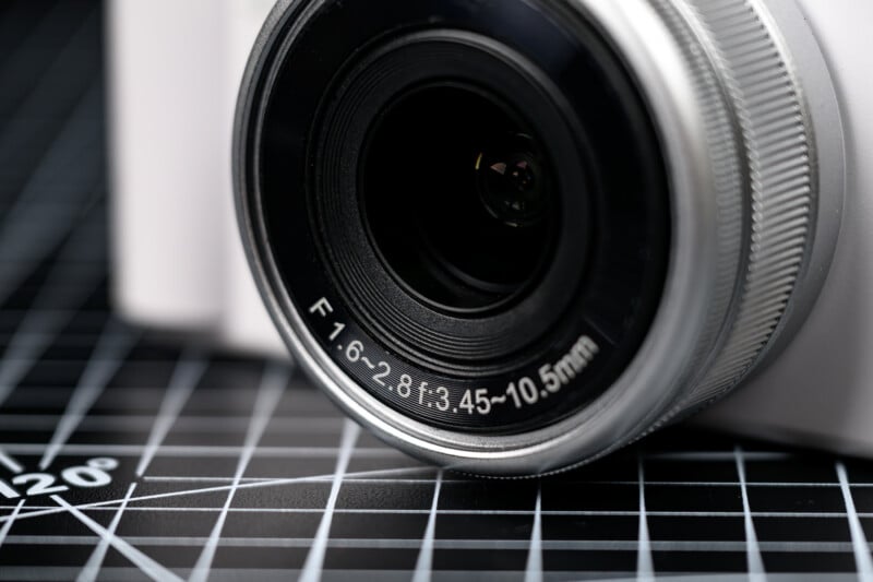 Close-up of a camera lens with focal length and aperture markings, set on a black grid cutting mat with white measurement lines.