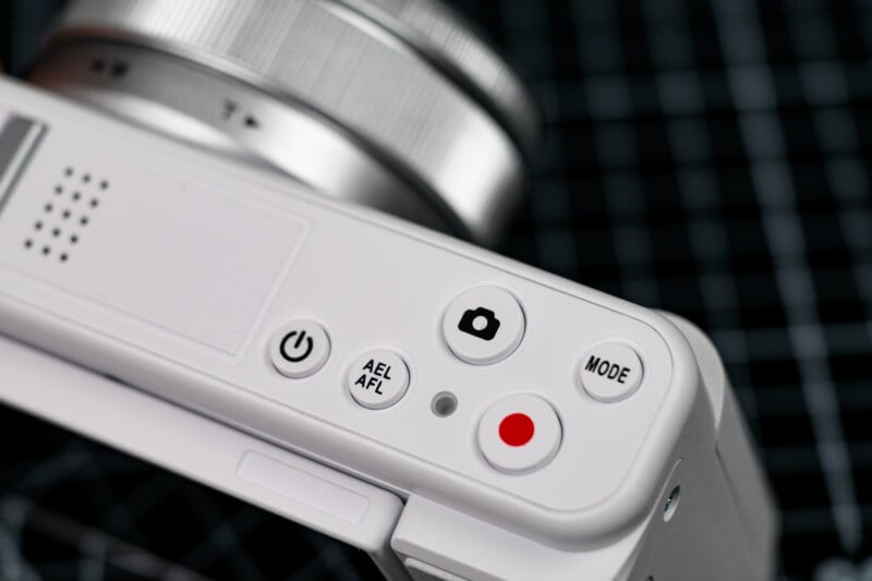 Close-up of a white digital camera showing buttons labeled power, AEL/AFL, a camera icon, mode, and a red record button, with part of the lens visible against a grid background.