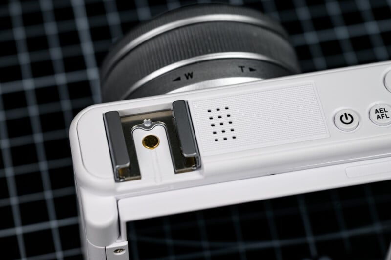 Close-up of a white digital camera’s top section showing the lens, power button, hot shoe mount, and part of a speaker grill, against a black grid-patterned background.