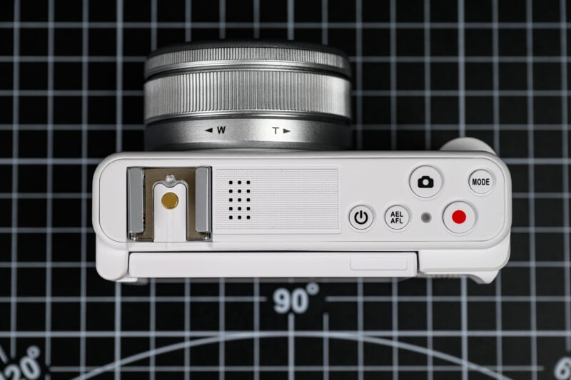 A silver and white digital camera viewed from above, showing its lens, control buttons, and dials, placed on a black grid background marked with degrees and measurement lines.