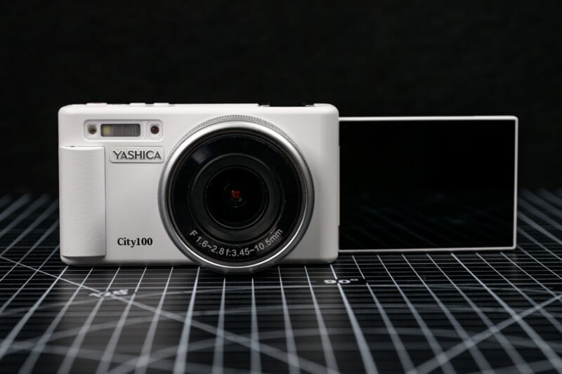 A white Yashica City100 digital camera with a flip-out LCD screen sits on a black grid-patterned surface against a dark background.