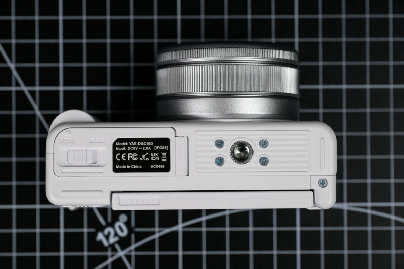 A close-up view of the bottom of a white digital camera with a silver lens, showing the tripod mount, battery compartment, and product label, placed on a black grid-patterned background.