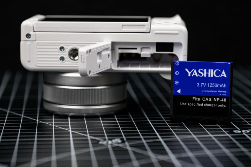 A close-up of a white digital camera with its battery compartment open, showing the slot. Next to it is a blue YASHICA 3.7V 1250mAh lithium polymer battery standing upright on a black grid surface.