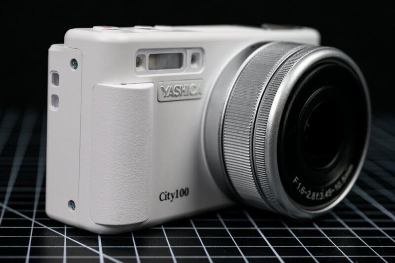A close-up view of a white compact digital camera labeled "Yashica City100," placed on a black grid-patterned surface. The lens is prominently featured on the right side of the image.