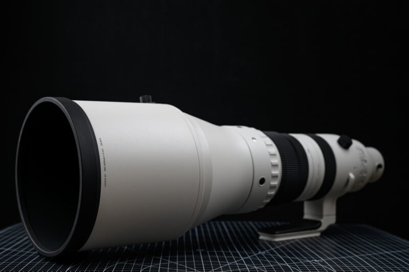 A large, white telephoto camera lens with a black rubber front ring, placed on a grid-patterned surface against a dark background.