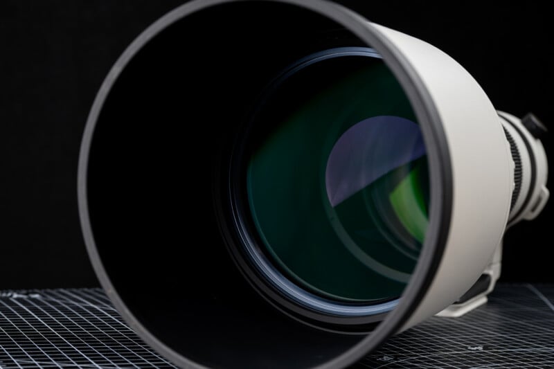Close-up of a large camera lens with a black lens hood, showing green reflections on the glass element, set against a dark background and a grid-patterned surface.