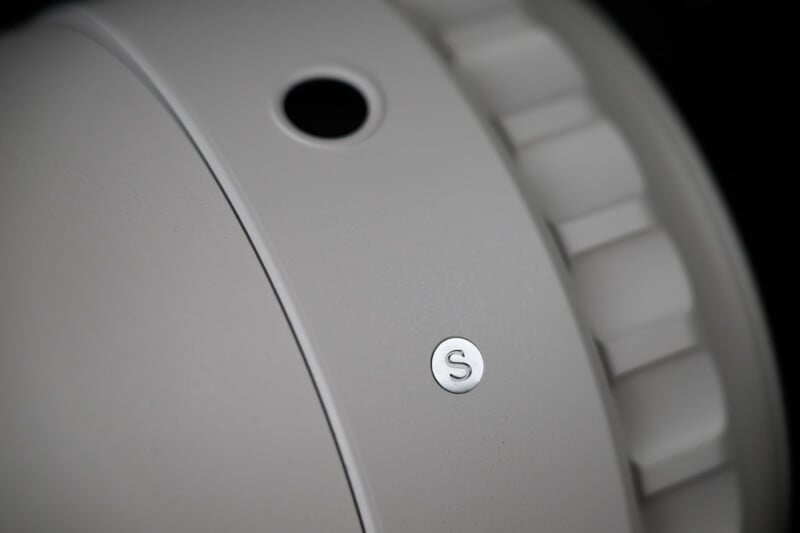 Close-up of a gray, round device with a small black hole, a textured dial, and a metallic button labeled "S" on its smooth surface.