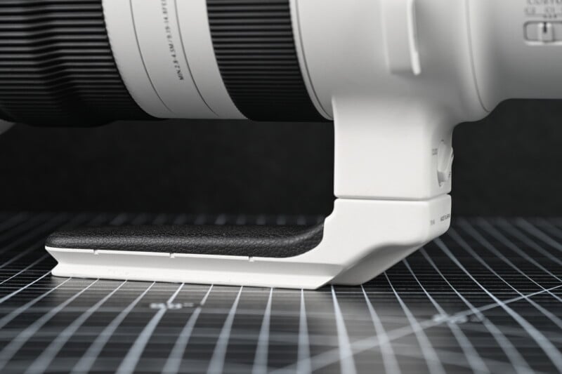 Close-up of a white camera lens with a black, textured tripod mount foot resting on a grid-patterned surface. The background is dark, making the lens and mount stand out.