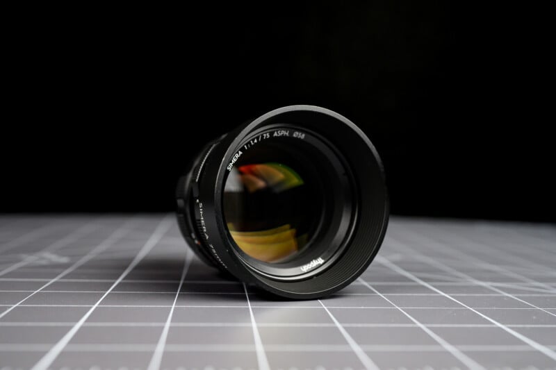 A camera lens positioned upright on a gray grid-patterned surface, with a dark blurred background. The focus is on the lens glass and markings around the rim.