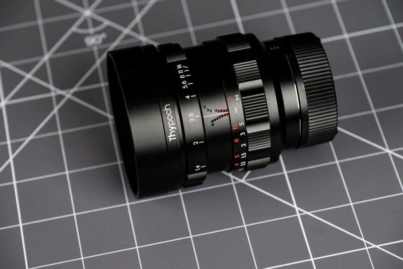 A black Thypoch camera lens with aperture and focus markings lies on a gray grid cutting mat.