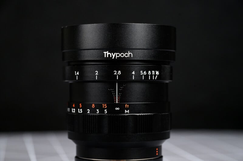 A close-up of a black Thypoch camera lens, showing aperture and focus markings in white and orange, set against a dark blurred background with a faint grid pattern below.