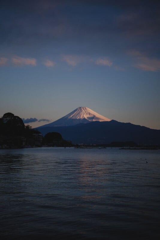 Mount Fuji covered in snow is illuminated by the setting sun, with a calm lake in the foreground and a dark silhouette of trees and buildings along the water’s edge. The sky is partly cloudy with warm, soft light.