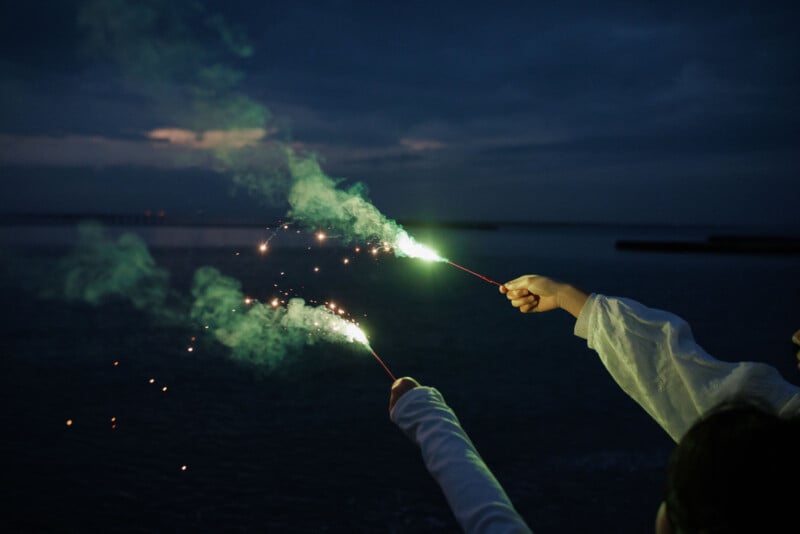 Two people hold sparklers, creating bright sparks and smoke against a dark, twilight sky by the water. Their hands and arms are visible, while the background is softly lit with a fading sunset.