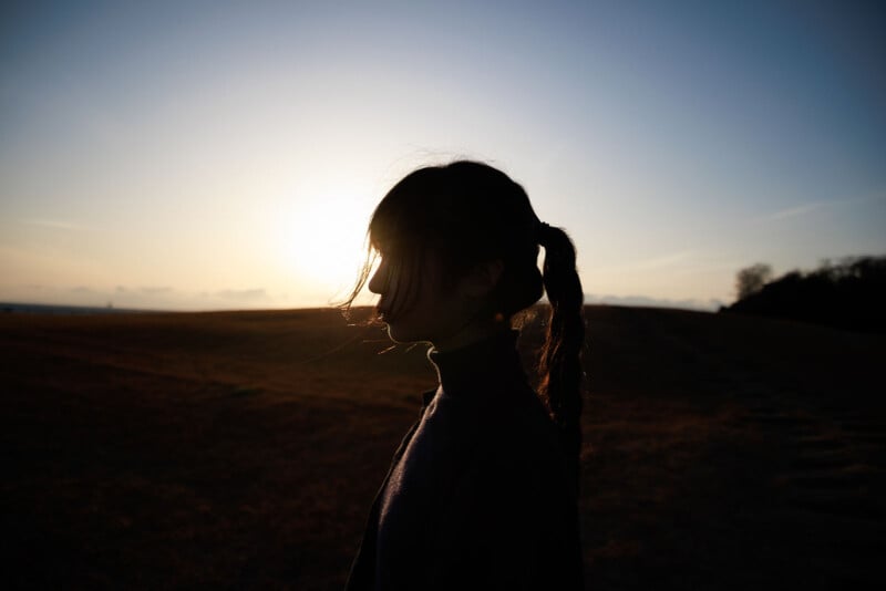 Silhouette of a person with a ponytail standing outdoors at sunset, with the sun low in the sky and casting a warm glow over an open field in the background.