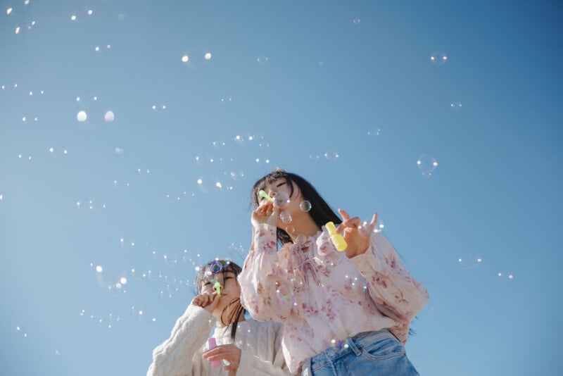 Two people stand outdoors under a clear blue sky, blowing bubbles. One wears a pink floral blouse and jeans, and the other wears a white sweater. Bubbles float around them, and both look relaxed and happy.