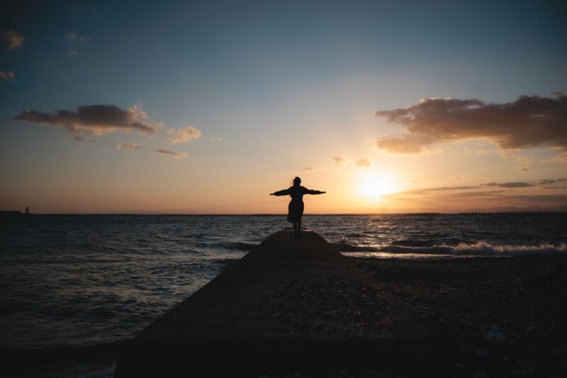 A person stands with arms outstretched at the end of a pier facing the ocean during sunset, silhouetted against a colorful sky with scattered clouds. Waves gently crash against the shore.